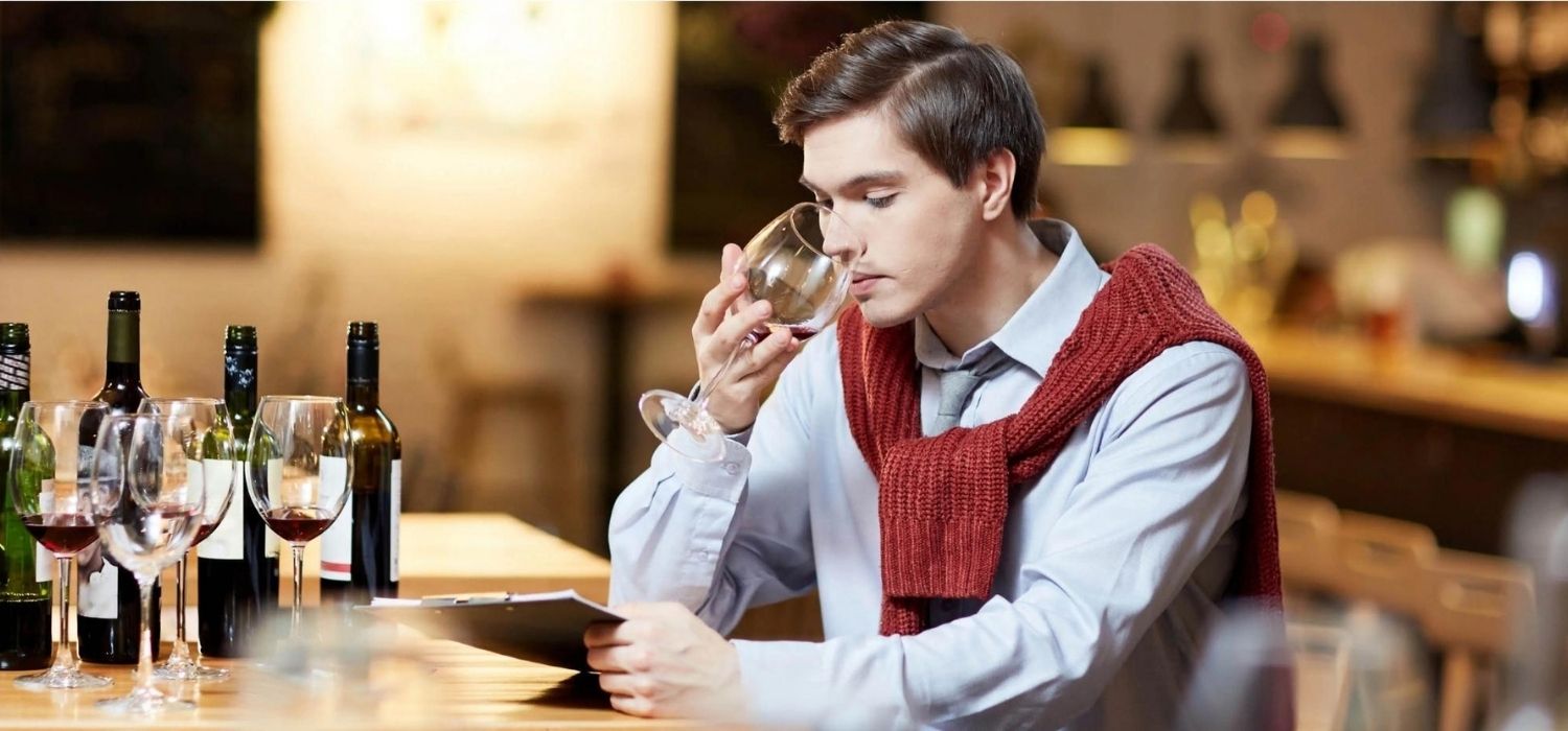 Un professionnel du vin évalue la qualité d’un vin rouge en le dégustant dans un bar ou une cave, entouré de bouteilles et de verres, illustrant le travail de sélection et de négociation dans le commerce du vin. - BSB